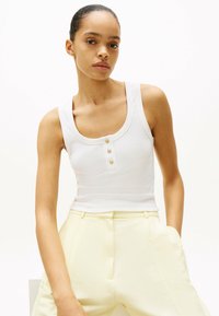 Top in cotone bianco a costine con scollatura a barca e tre bottoni dorati sul davanti. Il modello indossa pantaloni giallo chiaro sartoriali.