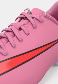 Vaaleanpunainen jalkapallokengä, jossa on sileä synteettinen päällinen, mustat yksityiskohdat ja suuri punainen swoosh-logo. Teksturoitu nauhoitus ja vaaleansininen pohja.