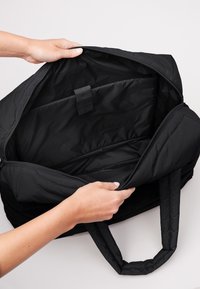 Händer som öppnar en svart vadderad tote-väska som visar ett rymligt fack och en vadderad laptopficka med rem inuti.