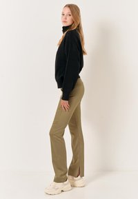 Schwarzer Strickpullover mit hohem Kragen, olivgrüne, taillierte Hose mit Seitenschlitzen und hellen, klobigen Sneakern. Glatte Texturen durchgehend.