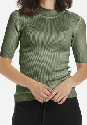 Femme portant un pull vert olive ajusté à manches courtes côtelées et un pantalon noir, les mains jointes à la taille.
