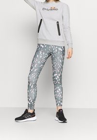 Sudadera gris con bolsillos con cremallera y logo. Leggings impresos en verde con patrones abstractos en rosa y blanco. Zapatillas deportivas negras.