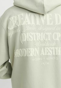 Hellgrüner Hoodie mit erhabenem weißen Text in verschiedenen Schriftarten. Das Design enthält Phrasen wie "Kreatives Viertel" und "Moderne Ästhetik."