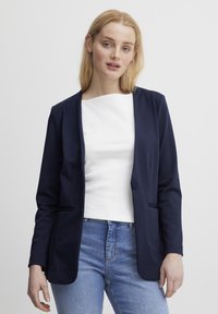 Námornícky modrý blazer vyrobený z mäkkej tkaniny, s klopovým golierom, jednobodovým zapínaním a dvoma prednými vreckami. Nosený cez biely top.