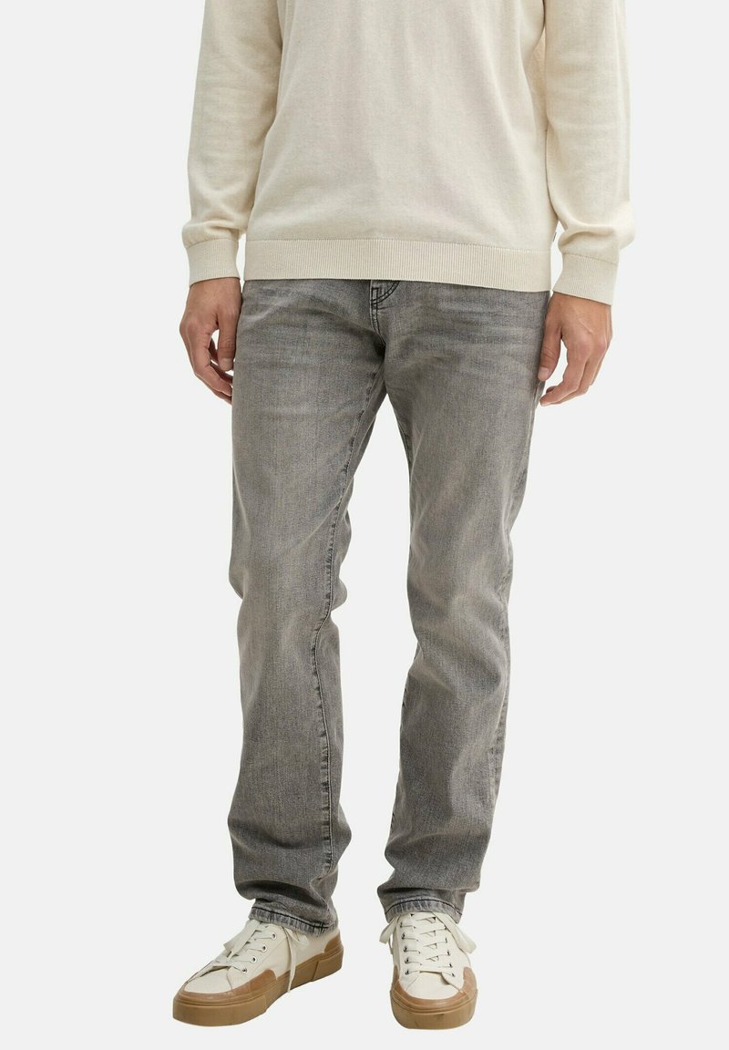 Szürke, slim fit farmernadrág, amely finom textúrával rendelkezik. Alacsony szárú, fehér sportcipővel párosítva, bézs részletekkel.