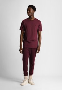 T-shirt bordeaux con collo a giro e logo discreto, abbinato a pantaloni della tuta bordeaux coordinati e sneakers beige; tessuto strutturato e vestibilità rilassata.