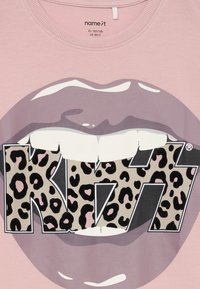 Rausvas marškinėliai su didelėmis violetinėmis lūpomis, kandžiojančiomis leopardų rašto "KISS" logotipą blokuotomis raidėmis per priekį.