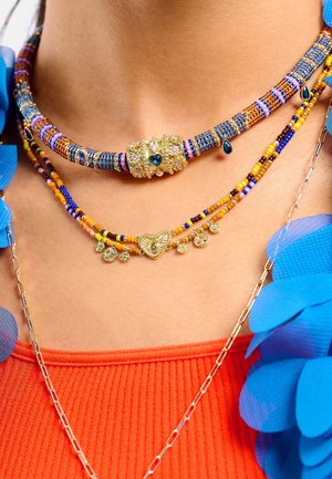 Trois colliers superposés avec des perles colorées en bleu, jaune et orange, ornés d'un pendentif en forme de cœur en or et d'accents décoratifs en métal.