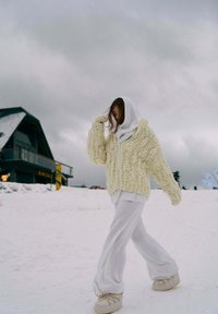 Žena v bielych nohaviciach, hrubom krémovom svetri s kapucňou kráča po snehu pri drevenej chate pod oblačnou oblohou.