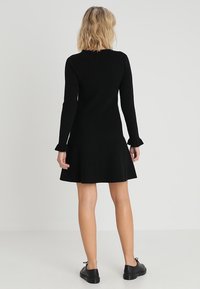 Abito nero a maglia con maniche lunghe, orlo svasato e polsini arricciati. Texture liscia, corpetto attillato e lunghezza fino a metà coscia.