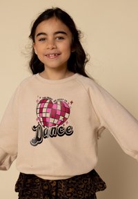 Lachend meisje met lang, donker haar, gekleed in een beige sweatshirt met een roze geruite hart en de tekst "Je laat me willen dansen."