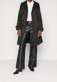 Mörkgrön trenchcoat med dubbelknapprad, tillsammans med en vit skjorta, svarta läderbyxor och klackstövlar. Släta texturer genomgående.