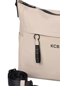 Bolso bandolera de color beige claro hecho de material sintético suave. Cuenta con un zipper negro, detalle del logo y una correa negra removible.