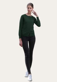 Cardigan vert avec un col rond, huit boutons et deux poches avant, associé à un jean skinny noir et des baskets blanches.