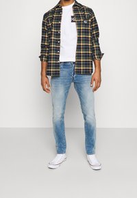 Person som bär blåa urtvättade jeans, vita högstukna sneakers, vit t-shirt och en svart och gul rutig skjorta med uppvikta ärmar, som är öppen.