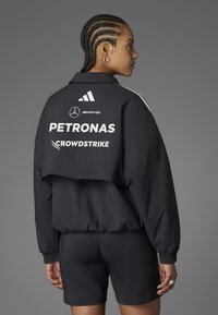 Crni kratak sako s bijelim logotipima, koji uključuje "PETRONAS" i "CROWDSTRIKE". Tekstuirana tkanina i kratki rukavi, u kombinaciji s odgovarajućim šeširima.