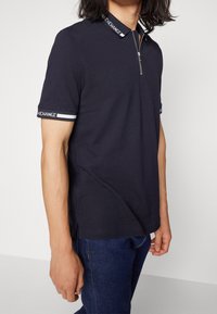 Polo-shirt navy à manches courtes avec un col zippé, présentant une bande de col contrastante et des accents de poignets rayés. Texture lisse, design ajusté.