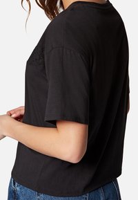 Černé zkrácené tričko s krátkým rukávem, které má texturované detaily na rameni. Vyrobeno z měkké bavlny. Hladký zadní díl a zaoblený lem.