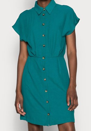 Robe chemise courte à manches courtes de couleur sarcelle, avec boutons sur le devant, col et taille cintrée, portée par une personne debout.