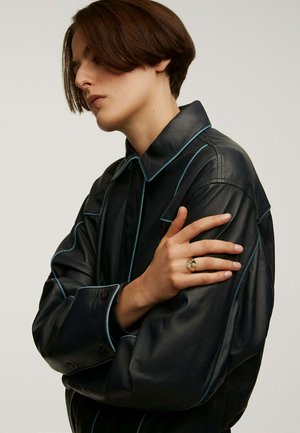 Chaqueta de cuero negra con costuras en contraste azul claro, gran cuello y cierres de botón. Una mano con un anillo dorado reposa sobre la manga.