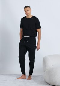 Νεαρός άνδρας, όρθιος ξυπόλητος σε χαλιάδα, φοράει μαύρο μπλουζάκι και μαύρο αθλητικό παντελόνι με λευκή επένδυση δίπλα σε λευκή επενδυμένη καρέκλα.