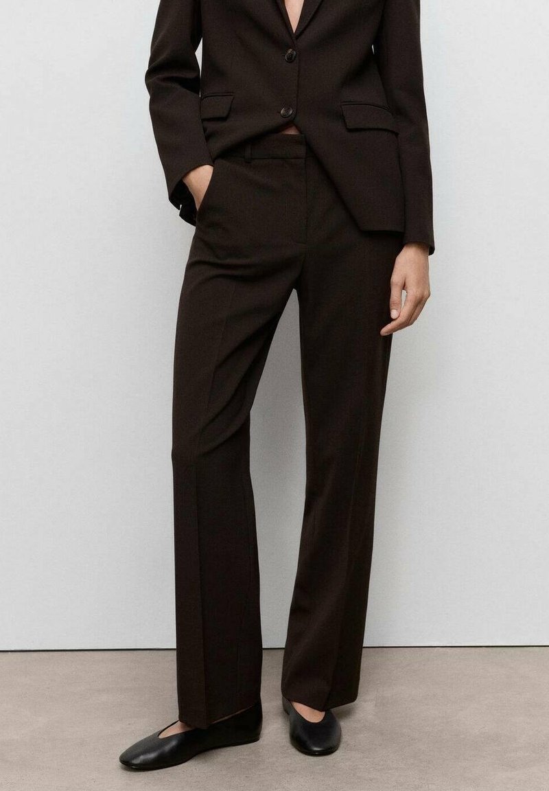 Femme portant un costume noir sur mesure avec un pantalon droit et des chaussures plates noires, debout contre un mur uni gris clair.