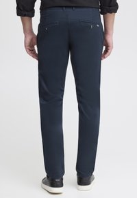 Muž stojící zády, oblečený v tmavě modrých úzkých kalhotách se zapínacími zadními kapsami a černými teniskami s bílou podešví.