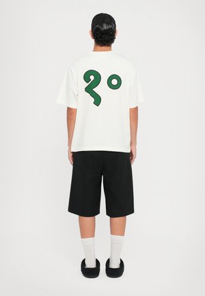 White t-shirt with a large green graphic of "2°" on the back, paired with black shorts and dark slip-on shoes, worn with white socks.