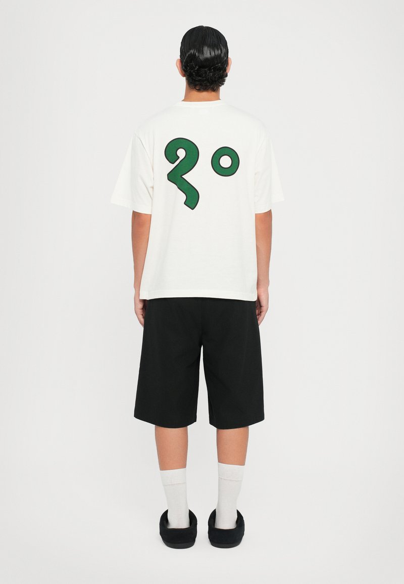 T-shirt branco com um grande gráfico verde de "2°" nas costas, combinado com calções pretos e sapatos escuros tipo slip-on, usados com meias brancas.