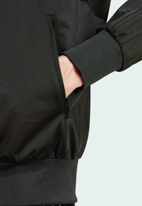 Zwarte sportjacket met een gestructureerde stof, een ritssluiting aan de zijkant en geribbelde manchetten. Het ontwerp benadrukt een gestroomlijnde, getailleerde silhouet.