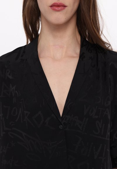 Svart blus med en mjuk textur och ett subtilt, tonalt graffiti-mönster. Den har en V-ringning och en enda svart knapppunkt.