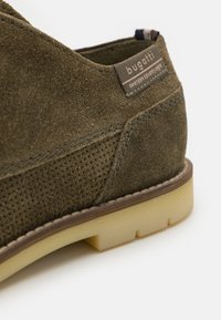 Zapato de ante verde oliva con detalles perforados, un acento de costura marrón y una suela de goma transparente con un acabado texturizado. Etiqueta de la marca visible.