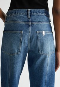 Jeans de mezclilla azul con cintura alta, pierna recta, costura en contraste y dos bolsillos traseros. Accesorios de metal en la cinturilla y el bolsillo.