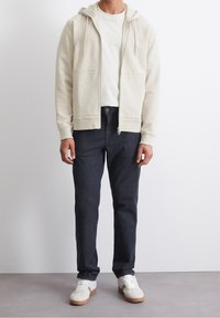 Felpa con zip color beige chiaro, dotata di cappuccio, abbinata a jeans scuri e scarpe da ginnastica bianche, in piedi su uno sfondo grigio. Design minimalista.