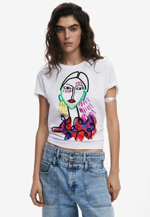 T-shirt en coton blanc avec un design de visage abstrait en noir, rouge, vert et violet. Style avec des manches retroussées et associé à un jean en denim bleu clair.