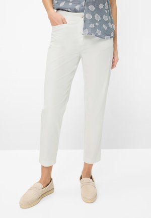 Kvinde iført hvide korte bukser, lyse beige slip-on sko og en blå blomstret skjorte med den ene hånd i lommen.