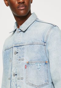 Chaqueta de mezclilla azul claro con un collar clásico, bolsillo frontal, detalles desgastados, costuras en contraste y botones metálicos.