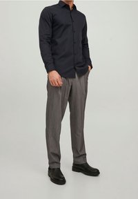 Camisa oscura de botones con mangas largas, combinada con pantalones de raya diplomática de color gris claro y botines negros. Ajuste nítido y a medida en todo el conjunto.