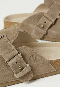 Brown suede slip-on sandals featuring a wide strap, adjustable buckle, textured insole, and a cork base. Minimalist design with embossed logo.
