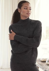 Maglione a collo alto grigio scuro realizzato in materiale morbido, con polsini e orlo a coste. Il modello posa con le braccia incrociate, mostrando la texture e la vestibilità.