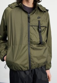 Nike lättviktsjacka med huva i olivgrön. Har dragkedja fram, bröstficka och justerbar dragsko vid fållen. Slät textur.