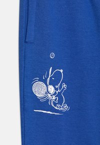 Modrá látka s bielym obrysom grafiky rozprávkového psa, ktorý hrá tenis, drží raketu a loptu nad hlavou. Textúra je jemná a hladká.