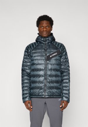 Gevoerde jas in donkerblauw met een ritssluiting, hoge kraag en een zwart logo patch op de borst. De stof heeft een glanzende afwerking en een gewatteerd ontwerp.