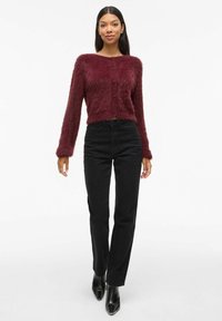Cardigan court en velours bordeaux avec des manches longues et des boutons sur le devant, associé à un jean noir taille haute et des bottines noires brillantes.
