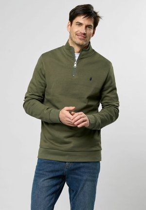 Olijfgroene ritsvest met een staande kraag, ribgebreide boorden en zoom, en een klein logo op de borst, gecombineerd met blauwe jeans.