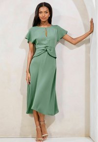 Vestito midi verde realizzato in tessuto morbido, con maniche a pipistrello corte, un'apertura a chiave frontale e un dettaglio annodato in vita. Scarpe con cinturini.
