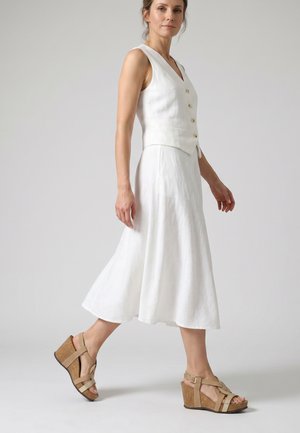 Woman walking, wearing a sleeveless white buttoned vest, white midi skirt, and beige wedge sandals with ankle straps.