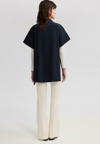 Top oversized din bumbac de culoare navy, cu mâneci scurte și guler, purtat peste o cămașă albă cu mânecă lungă și pantaloni evazați de culoare crem.