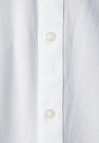 Camicia di cotone bianca con una fila di quattro bottoni, tessuto testurizzato e un design di colletto pulito e classico. La vista ravvicinata mette in evidenza i dettagli della cucitura.