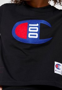 Černá mikina se širokým modrým, červeným a bílým logem s nápisem "100" a malou cedulkou v pravém dolním rohu. Měkký bavlněný materiál.